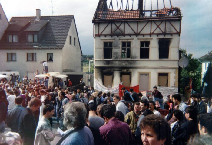 Ein frei stehendes Haus mit ausgebranntem Dachstuhl und verkohlten Fenstern, davor eine Menschenmenge