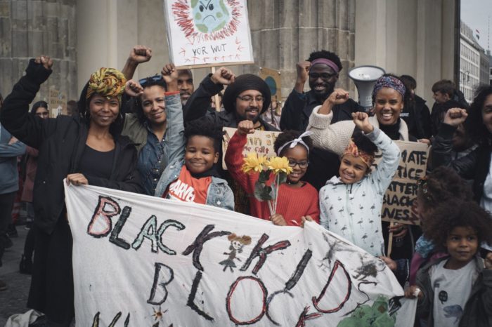 Erwachsene und Kinder heben die Black Power Faust vor einem Plakat mit der Aufsschrift Black Kids Block