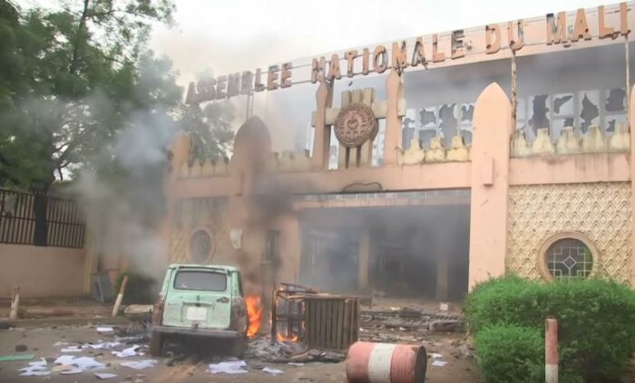 Eingang zum Parlamentsgebäude mit den Lettern Assemblee Nationale du Mali, die Lettern und die Glasfront sind beschädigt. Davor ein brennendes Auto und lose Blätter