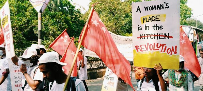 Demonstrantinnen und Demonstranten mit Flaggen, auf einem Plakat steht "A Woman's place is in the Kitchen", kitchen ist durchgestrichen und stattdessen steht revolution