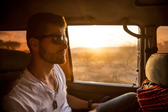 Weißer Mann mit Sonnenbrille blickt cool in einem Jeep vor einer Savannenlandschaft drein