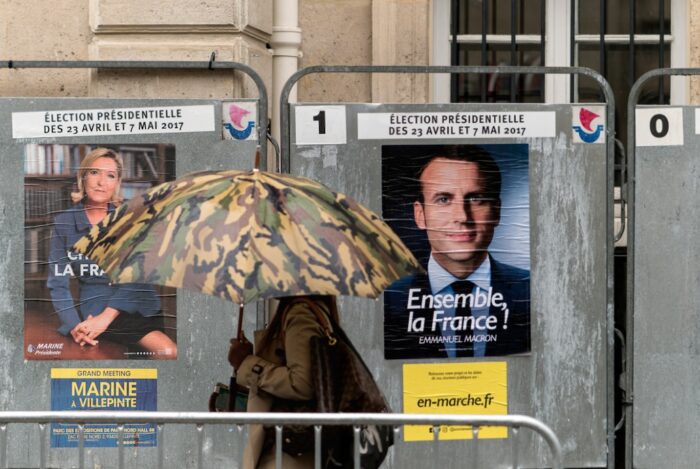 Eine Person mit Regenschirm geht an einer Wand vorbei, an der zwei Plakate hängen, eines mit Macron und eines mit Le Pen.