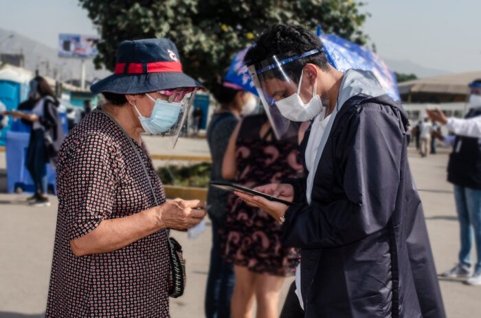 Eine etwa ältere Frau und ein jüngerer Mann, beide mit Masken und Schutzvisieren, schauen auf eine Wählerliste.