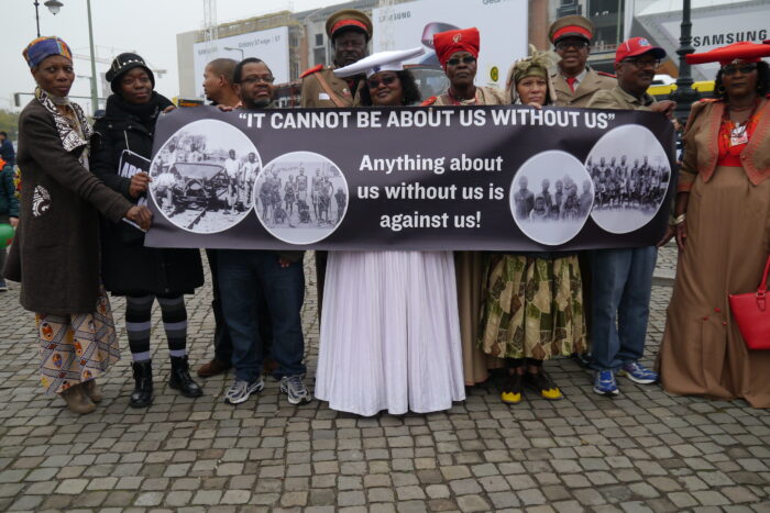 Vertreterinnen und vertreter der Herero und Nama halten ein Schild mit Archivbilder deutscher Verbrechen, wo drauf steht "It cannot be about us without us" und unten "anything about us without us is against us"