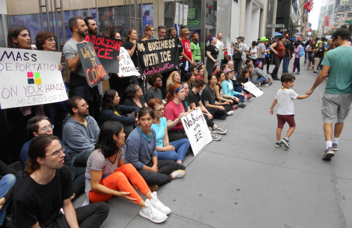 Demonstrantinnen und Demonstranten stehen und Sitzen vor einem Gebäude. Sie halten Schilder in der Hand, die die Zusammenarbeit von Microsoft und der Zollbehörde ICE verurteilen