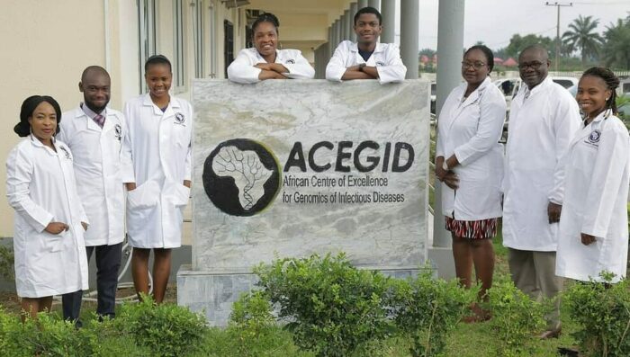 Ein Team von Forscher_innen im Familienfoto um eine Infotafel mit der Aufschrift "ACEGID, African Centre of Excellence for Genomics of Infectious Diseases"