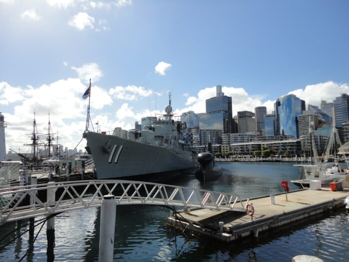 Pier mit einem Kriegsschiff und einem Uboot, im Hintergrund die Skyline von Sydney