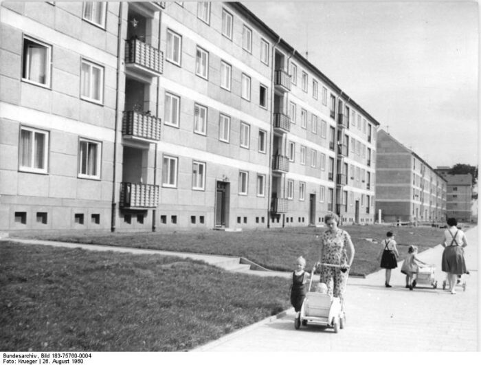 Schwarz-Weiß-Foto einer Häusersiedlung, auf dem Gehweg schiebt eine Frau einen Kinderwagen, ein kleines Kind läuft neben ihr, weitere Kinder dahinter
