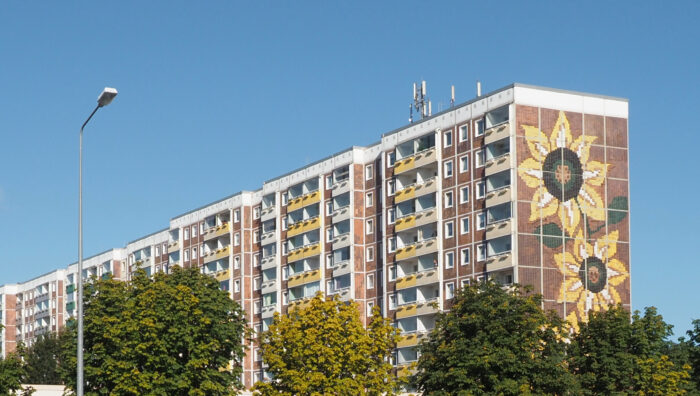 Bild des Sonnenblumenhauses, einem Plattenbau mit Balkonen. An der Seite prangt ein Meterhohes Bild von zwei Sonnenblumen. Der Himmel ist Blau.