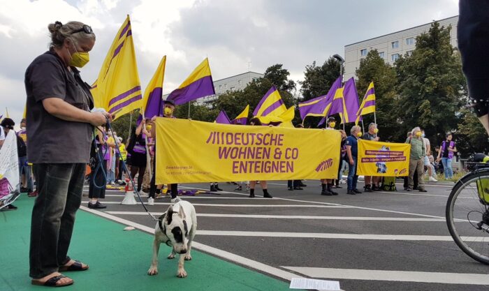 Demonstration mit Flaggen und Banner von Deutsche Wohnen und Co Enteignen. Links eine Person mit einem Hund an der Leine