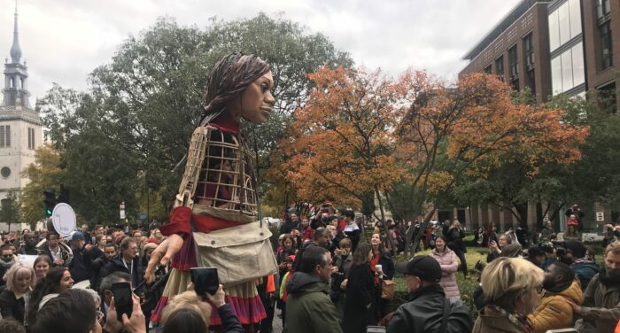 Eine Riesige, 3,5 Meter hoch, läuft durch eine Gruppe von Menschen, viele von ihnen fotographieren. Die Puppe sieht aus wie ein klines Mädchen. Im Brustkorb ist ein Mann zu sehen, der die Puppe steuert.