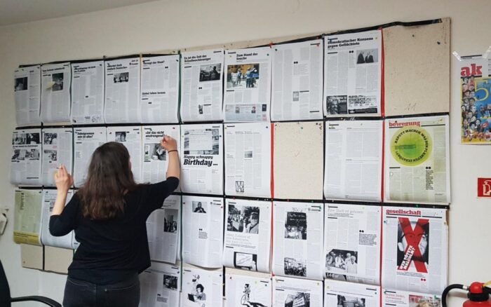 Eine rauchende Frau von hinten, steht vor einer Pinnwand, an der Zeitungsseiten hängen