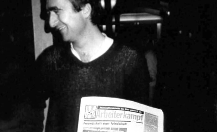 Schwarzweißaufnahme eines lachenden Mannes, der die Zeitung Arbeiterkampf in der Hand hält