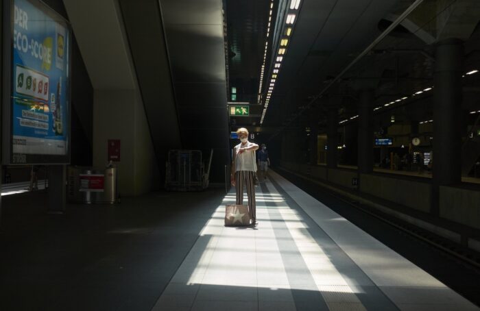 Eine Frau steht an einem Bahnsteig, die Umgebung ist sehr dunkel, die Frau steht in einem Lichtkegel und schaut auf die Uhr