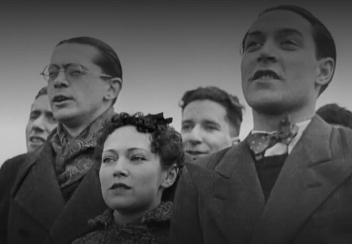 Auf dem Screenshot aus einem Schwarz-Weiß-Film sind vier nah beieinanderstehende Personen zu sehen, die aufrecht stehen und die Internationale singen.
