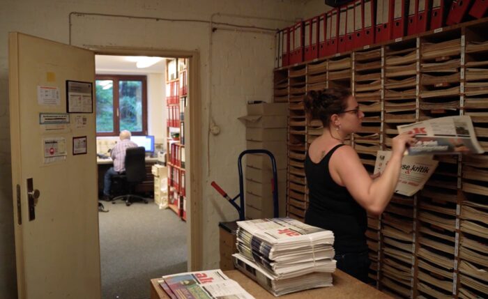 Foto: Ein düsterer Raum, Hannah Eberle sortiert Zeitungen in fächer, im Vordergrund liegt ein Zeitungsstapel auf dem Tisch, im Hintergrund steht eine Tür in einen anderen Raum offen, wo jemand am Computer sitzt