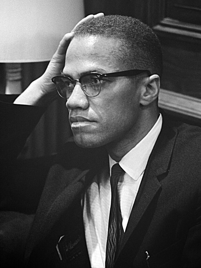 Porträt von Malcom X. Er scheint aufmerksam zuzuhören, während er seinen Kopf mit dem rechten Arm stützt.