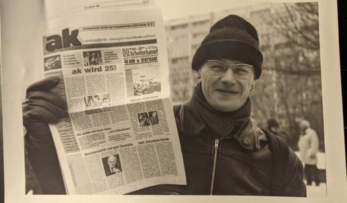 Schwarzweißaufnahme eines lächelnden Mannes mit Mütze, der eine Zeitung in die Höhe hält, Schlagzeile: ak wird 25