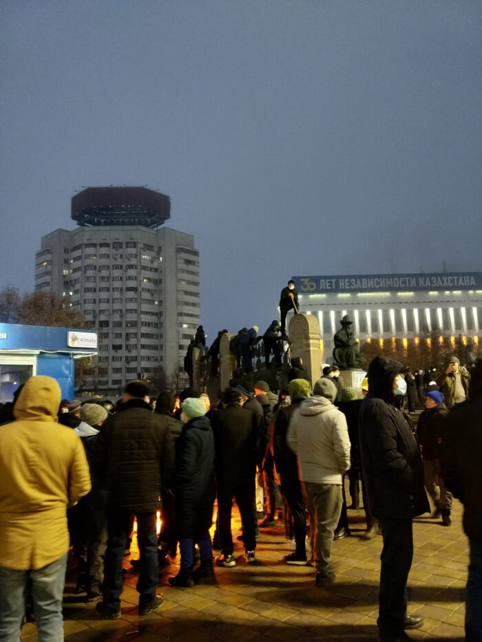 Nachtaufnahme einer Menschenmenge auf einem Platz in einer Stadt, an einer Reklame ist das Wort "Almaty" zu lesen