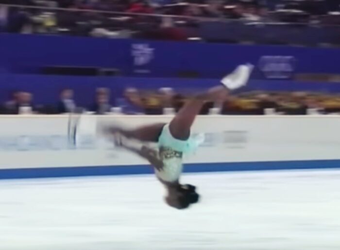 Man sieht eine Eiskunstläuferin mit dem Kopf nach unten und den Beinen nach oben, sie ist mitten in einem Salto rückwärts.