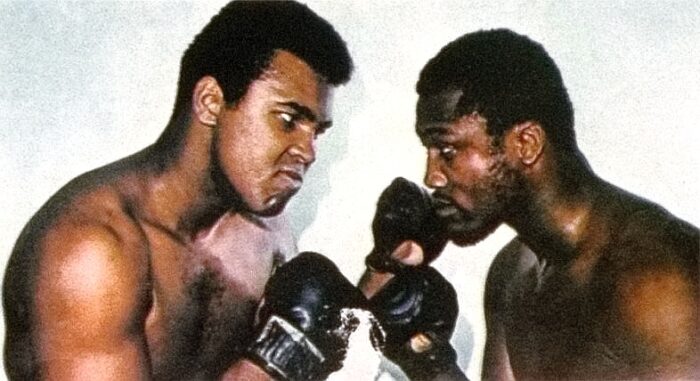 Ein gestelltes Box foto zeigt Muhammad Ali und Joe Frazier, die sich gegenüber stehen, einander böse anschauen und die Fäuste heben.