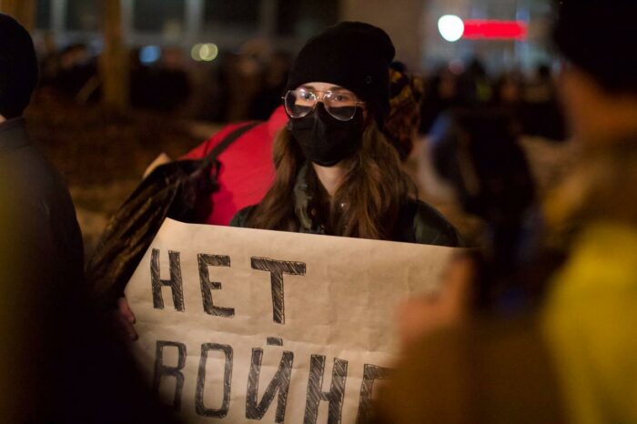Eine Person mit Maske und Schwarzer Mütze steht in einer Menge bei Nacht und hält ein Schild, wo in kyrillischer Schrift draufsteht "kein Krieg"