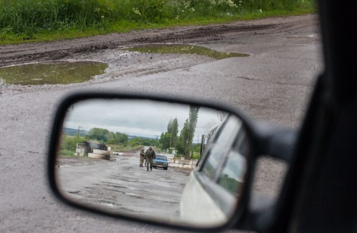 Im Rückspiegel eines Autos sieht man eine Straßensperre auf einer verregneten Landstraße, von der ein Mann in Uniform auf das Auto zugeht.