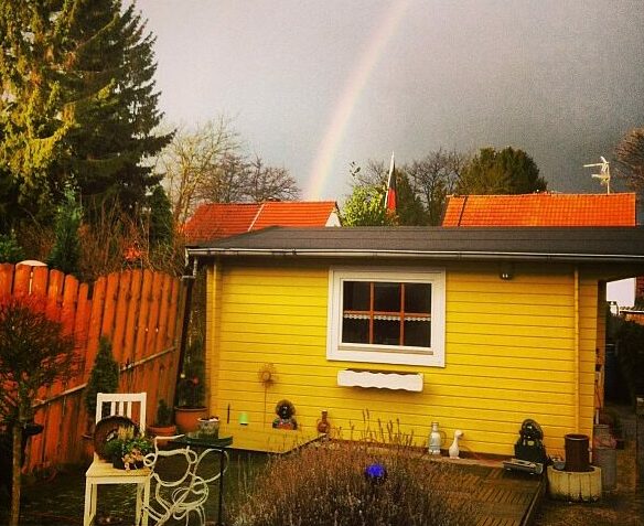 Das Foto zeigt ein gelbes Gartenhäuschen. Über dem Gartenhäuschen ist ein großer Regenbogen am Himmel zu sehen.
