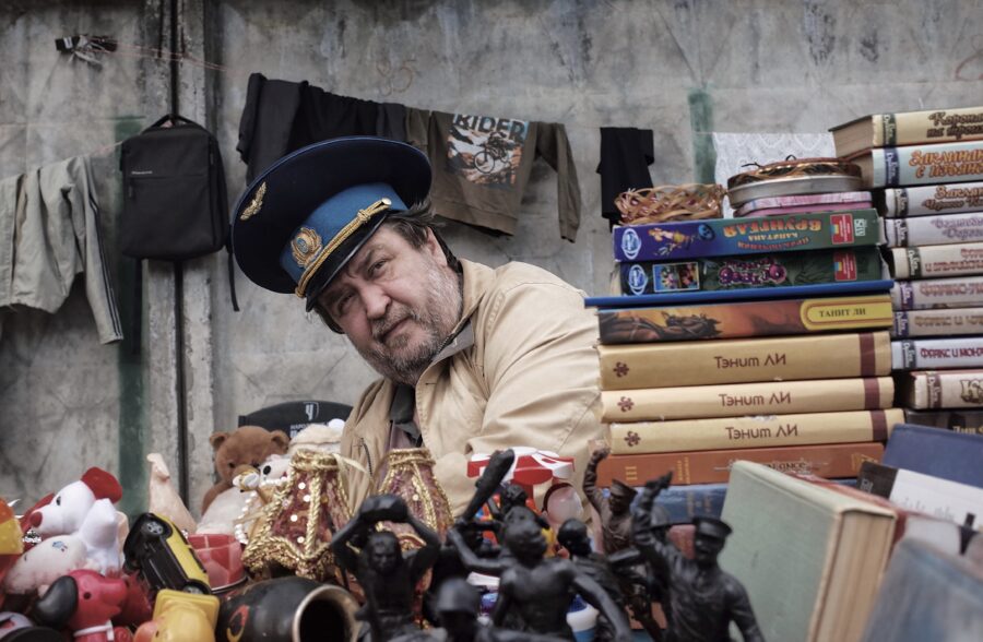 Ein Mann mit altertümlicher Militärmütze sitzt an einem Flohmarktstand hinter einem Haufen Figuren, Bücher, Videokassetten