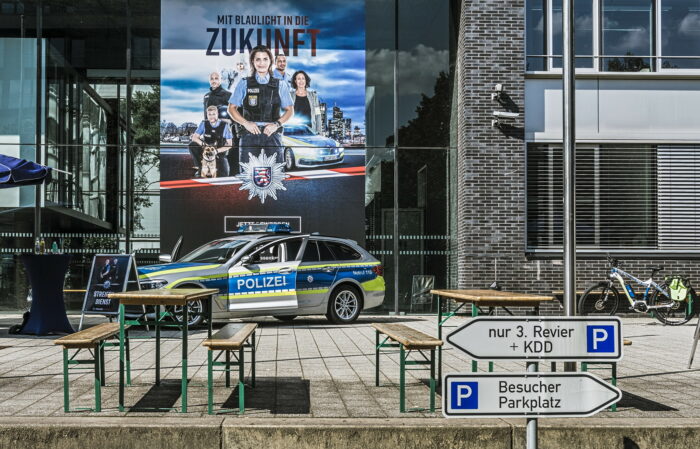 Das Foto zeigt ein Rekrutierungs-Event der hessischen Polizei vor einem Gebäude. Im Hintergrund hängt ein gigantisches Plakat von mindestens fünf Metern Höhe und drei Metern Breite. Auf dem Plakat posieren Polizist*innen und auch ein Polizeihund. Alle sehen sehr glücklich aus. Vor dem Plakat steht ein Polizei-Auto mit offenen Türen. Im Vordergrund des Bildes sind mehrere Bierbank-Garnituren zu sehen. Sie sollen wohl zum Verweilen einladen.