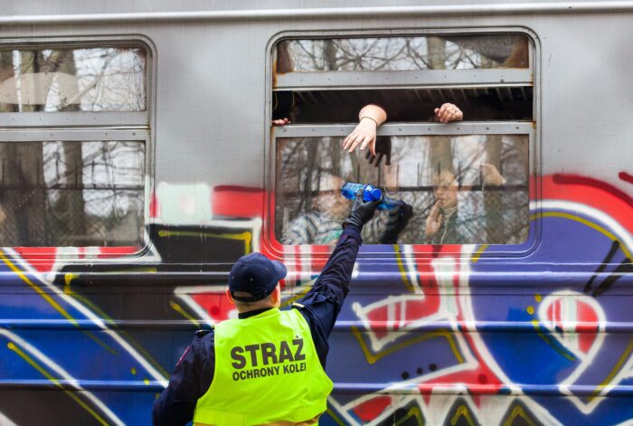Ein uniformierter Mann reicht eine Wasserflasche durch in ein Zugenster, ein Arm versucht aus dem Fenster, die Flasche zu greifen. Hinter der Scheibe im Zug sind undeutlich Kinder zu erkennen
