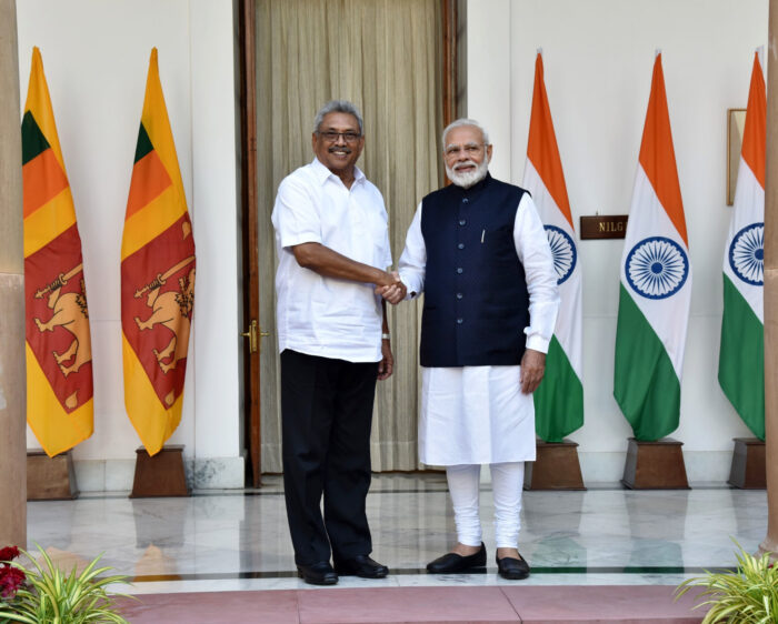 Zwei Staatsleute (siehe Foto-Beschreibung) geben sich die Hand und schauen in die Kamera. Im Hintergrund sind rechts drei Flaggenvon Indien, links drei Flaggen ovn Sri Lanka.