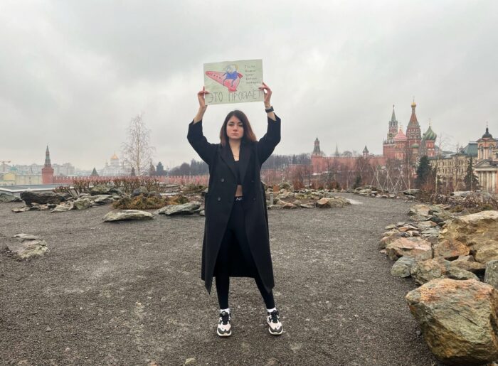 Eine junge Frau auf einem Schotterplatz hält ein Schild mit einer Putin-Karikatur und einer Aufschrift hoch, im Hintergrund die Mauern des Kreml und die Türme des Roten Platzes.