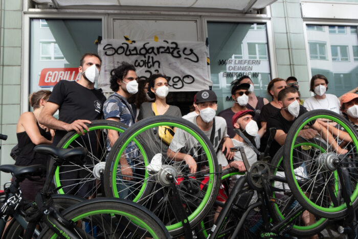 Auf dem Bild sind mehrere Menschen vor dem Eingang eines Warenlagers zu sehen. Vor ihnen stehen umgedrehte Fahrräder, hinter ihnen ein Banner, wo drauf steht "Solidarity with Santiago"