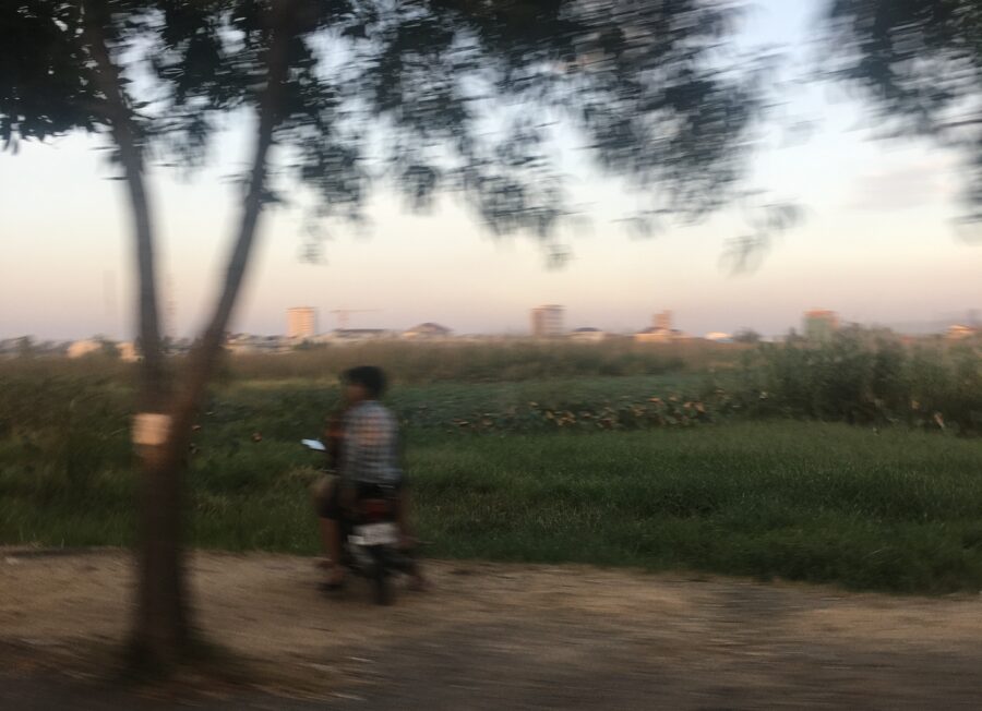 Verschwommene Aufnahme von zwei Menschen auf einem Motorroller am Straßenrand, links ein Baum, im Hintergrund ein Feld und ganz hinten schmenhaft Häuser