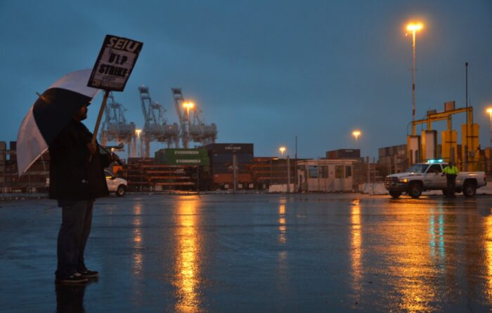 Ein Mann mit Regenschirm und Schild (Auschrift: SEIU ULP Strike) steht auf einem verregneten Hafenparkplatz, im Hintergrund Containerkräne und ein Auto
