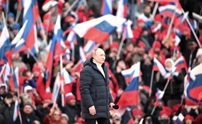 Vladimir Putin in winter coat with microphone in front of a crowd waving Russian flags