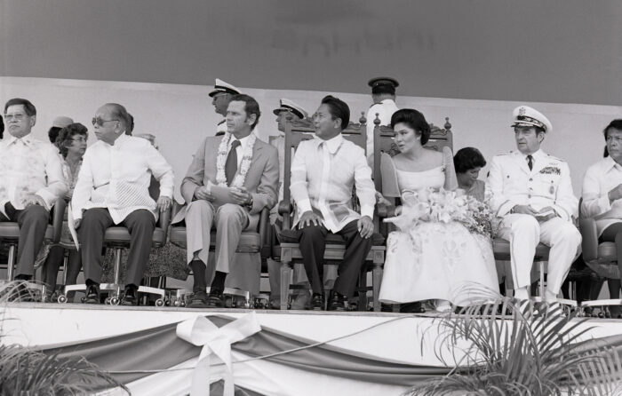 Mehrere Menschen sitzen bei einer Zeremonie auf einem Podium. Darunterauch Menschen in Militäruniform. Alle schauen weg von der Kamera auf ein Ereignis.