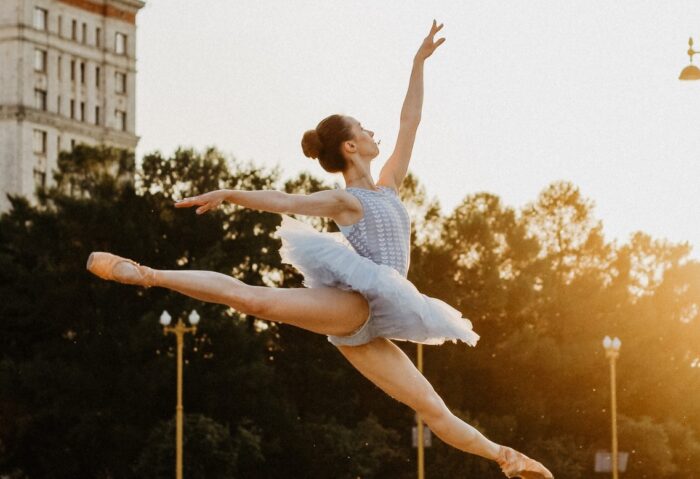 Man sieht eine Ballerina, die in der Luft einen Spagat macht, im Freien auf der Straße, im Hintergrund sind Häuser zu sehen.
