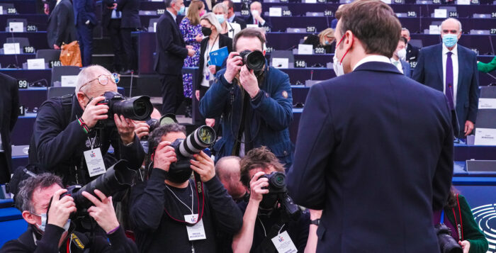 Bild aus einem Parlament. Die meisten Reihen sind leer. Vorne im Bild Macron mit dem Rücken zur Kamera. Vor ihm knien Fotographen und machen Bilder von ihm.