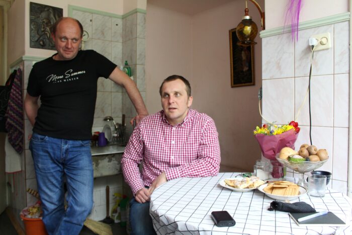 One man sitting at a table with flowers and biscuits on it, another mn leaning on the first man's chair and smiling