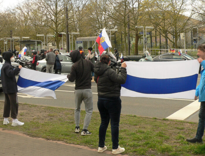 Bild einer Demonstration. Im Hintergrund eine Ansammlung von Menschen zwischen Autos. Einige halten russische Flaggen. Im Vordergrund vier Gegendemonstrant*innen, die zwei weiß-blau-weiße Flagge der russischen Opposition halten.