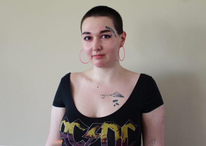 Ksenia vom Queer Lab ht kurze Haare und mehrere Tattoos, sie trägt ein AC/DC-Shirt und große Ohrringe.