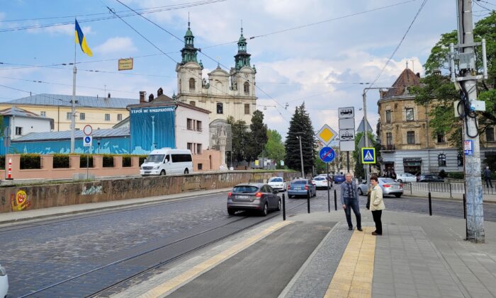 Eine Straße, hinten rechts stehen zwei ältere Menschen, hinten links ist eine Kirche zu sehen und ein Fahnenmast mit einer Landesflagge
