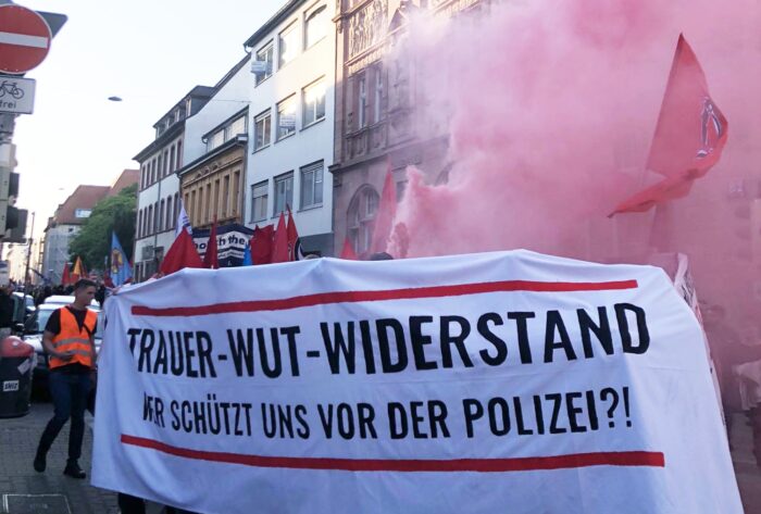 Eine Demonstration zieht duch die Straße, im Vordergrund ein Transparent mit der Aufschrift "Trauer - Wut - Widerstand: Wer schätzt uns vor der Polizei?", dahinter Rauch von einem Bengalo und eine Antifa-Fahne