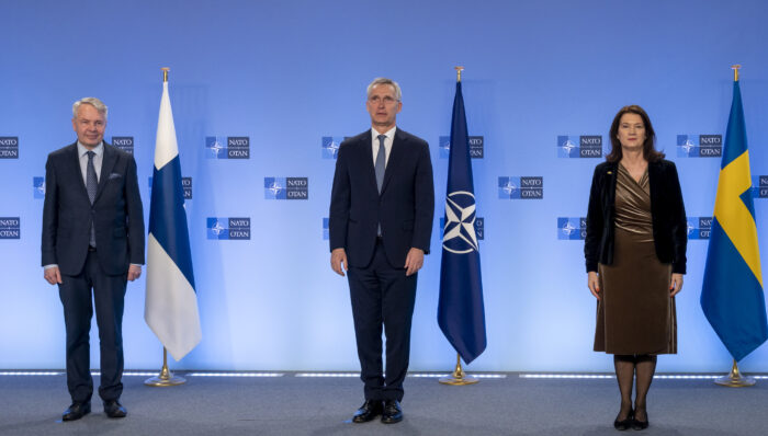 Ein Mann steht vor einer finnischen Flagge, ein anderer Mann vor einer Nato-Fahne und eine Frau vor einer schwedischen Fahne