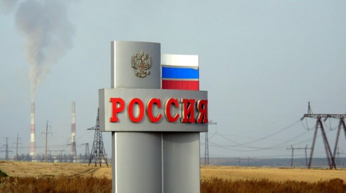 Ein großes Schild mit der Aufschrift "Russland" vor einer Steppenlandschaft, im Hintergrund sieht man Stromtrassen und rauchende Schonrsteine