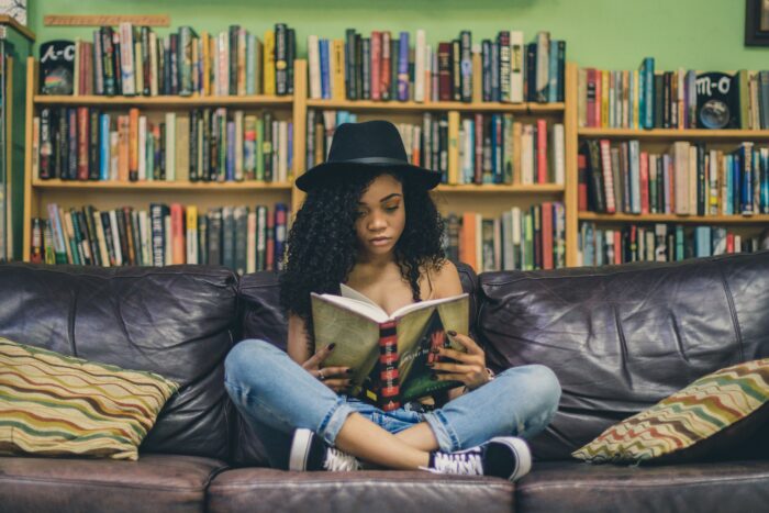 Bild einer Schwarzen Frau, die auf einer Couch sitzt und ein Buch liest. Hinter ihr ein großes Regal voller Bücher