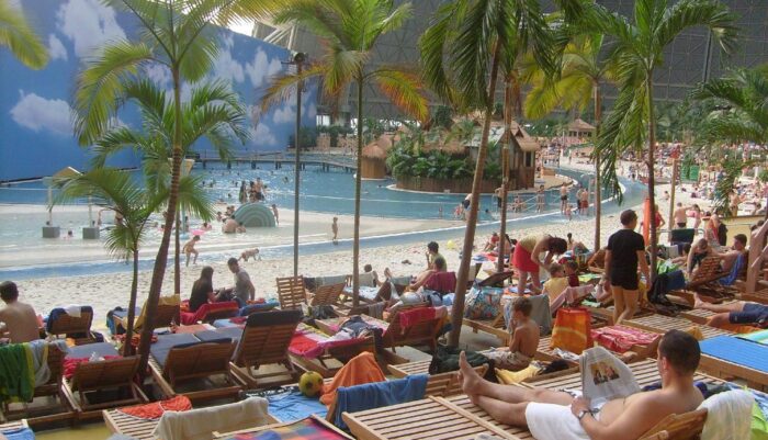 Ausschnitt aus tropical Island. Im Hintergrund umkreist eine metallische Wand die Halle, unterbrochen vor einem Fenster. Vor dem Fenster steht eine Wand mit Zeichnungen von Wolten. Es gibt einen Sandstrand und im Wasser plantschen Kinder. AStrand liegem Menschen auf Liegestühlen