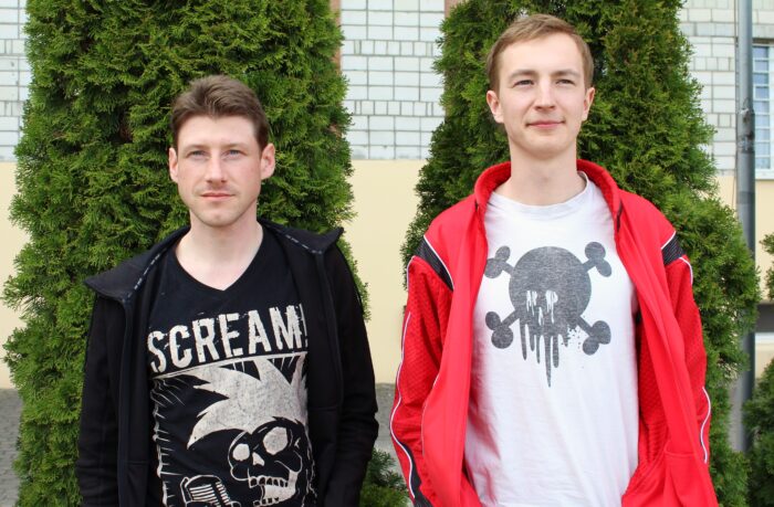Zwei junge Männer in Punk-Shirts stehen vor einer Hecke